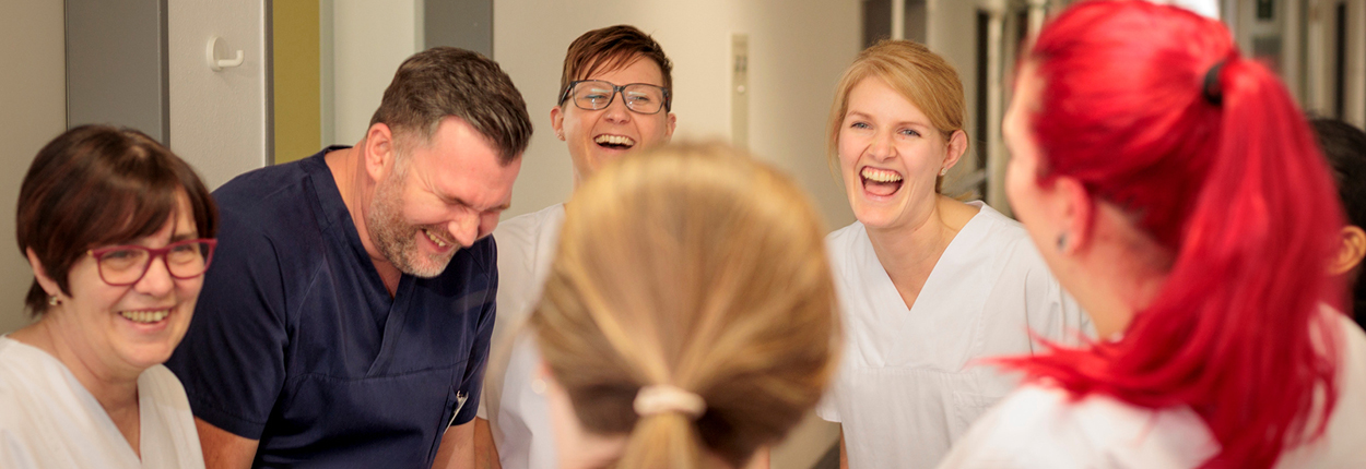 Gruppe von sechs medizinischen Fachkräften in weißer und blauer Arbeitskleidung, die gemeinsam lachen und sich unterhalten